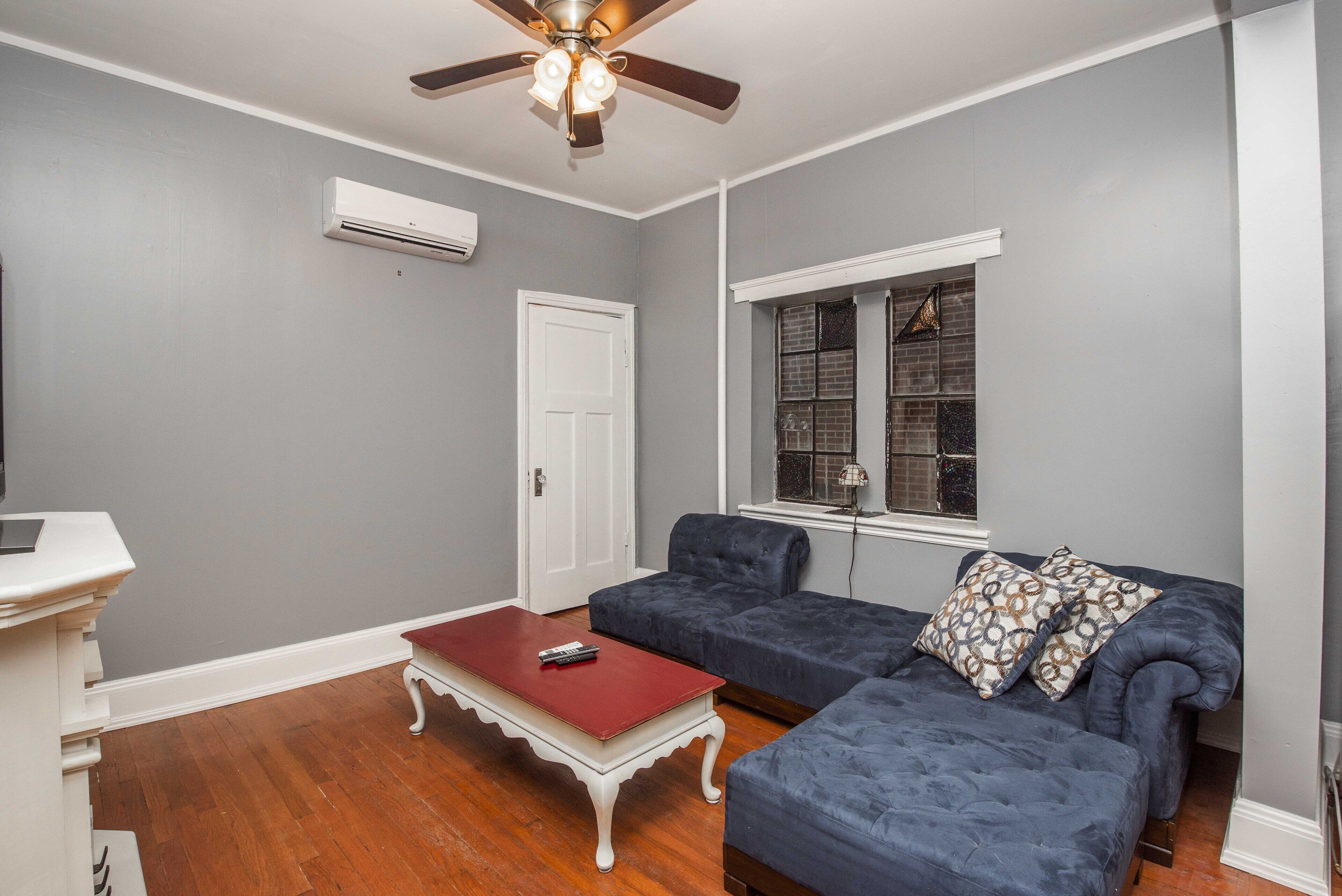 A living room with a ceiling fan, a fireplace, a white door, and a window with a view of the outside.