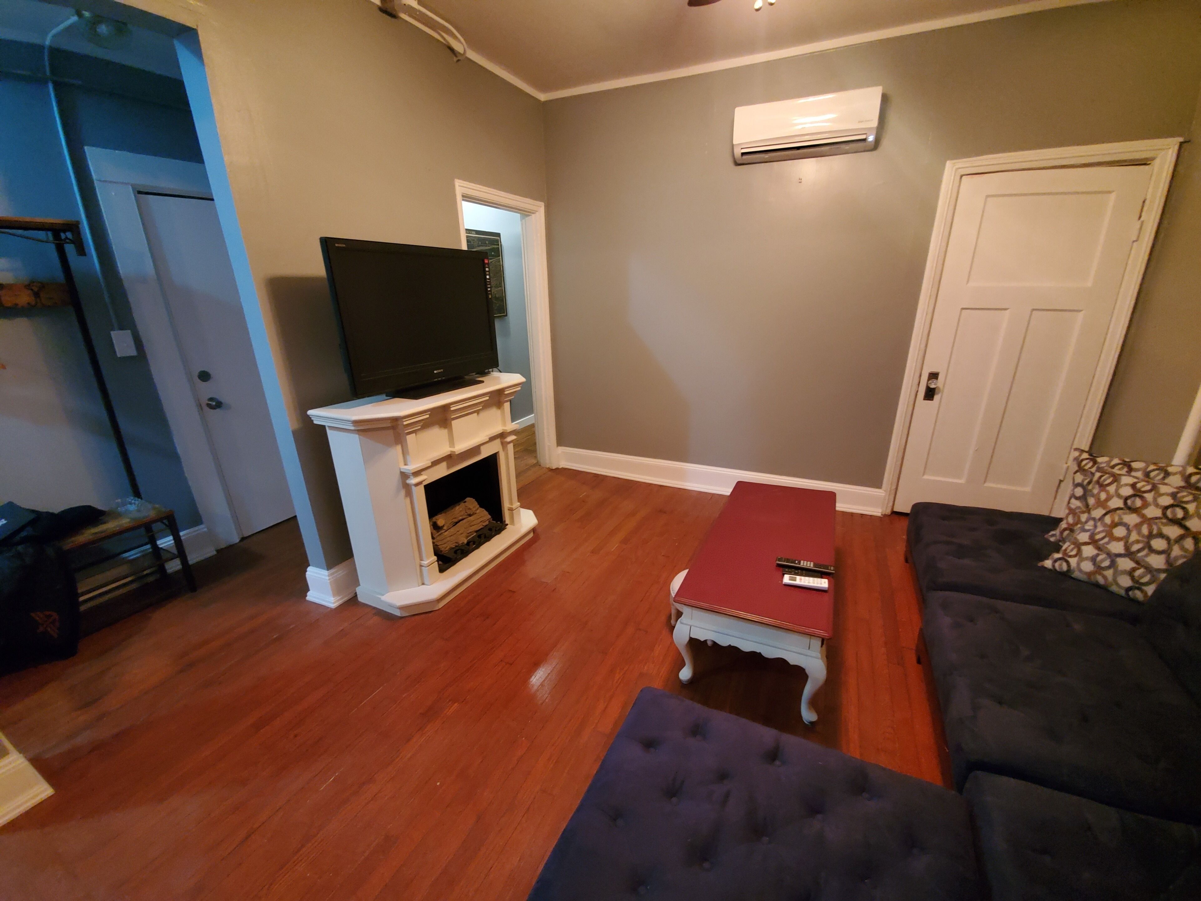 A living room with a fireplace, a television, a red ottoman, and a dark-colored sofa.
