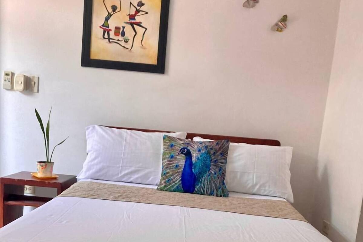 A neatly made bed with white linens and a blue peacock pillow, a small table with a plant, and a framed abstract painting on the wall.