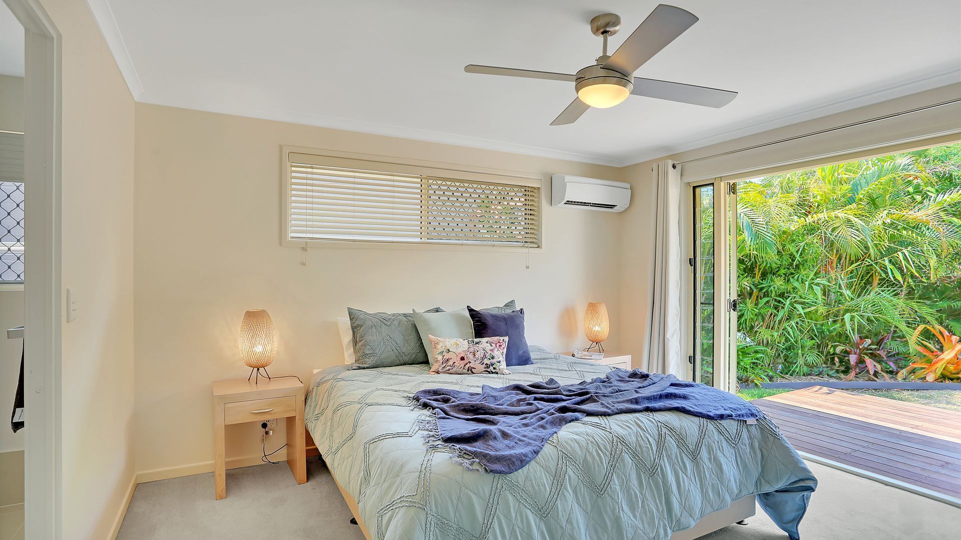 A bedroom with a bed, two bedside lamps, a ceiling fan, and a view of greenery through a sliding glass door.