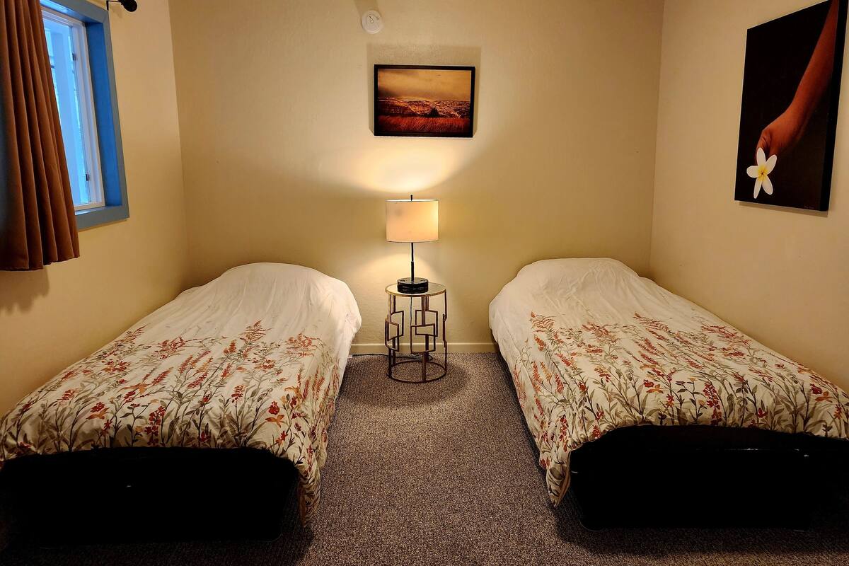A room with two single beds, a small table with a lamp, a painting on the wall, and a window with curtains.