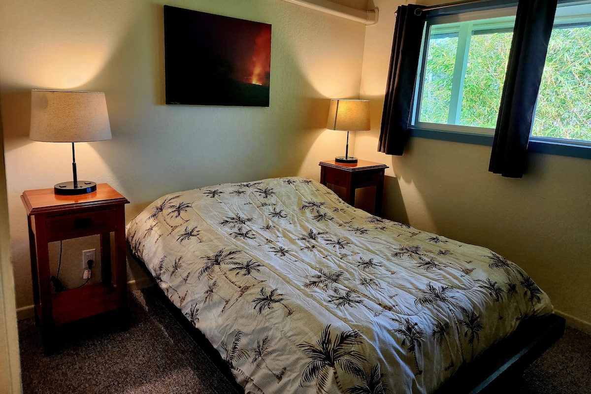 A bedroom with a bed, two bedside tables, a wall-mounted air conditioner, a window with curtains, and a painting on the wall.
