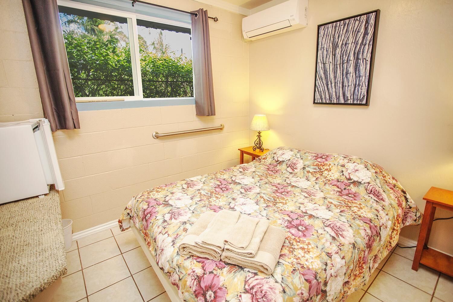 Un dormitorio con edredón estampado, lámpara de noche, radiador y ventana con cortinas.