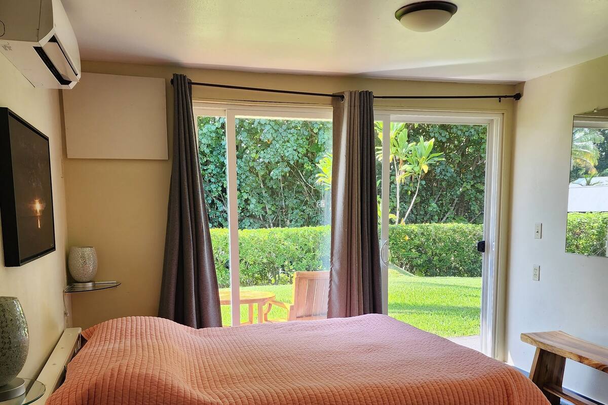 A bedroom with a large bed, a window with curtains, a bedside table, and a view of greenery outside.
