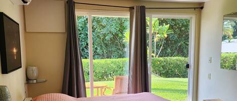 Quarto com cama grande, janela com cortinas, mesa de cabeceira e vista para o verde lá fora.