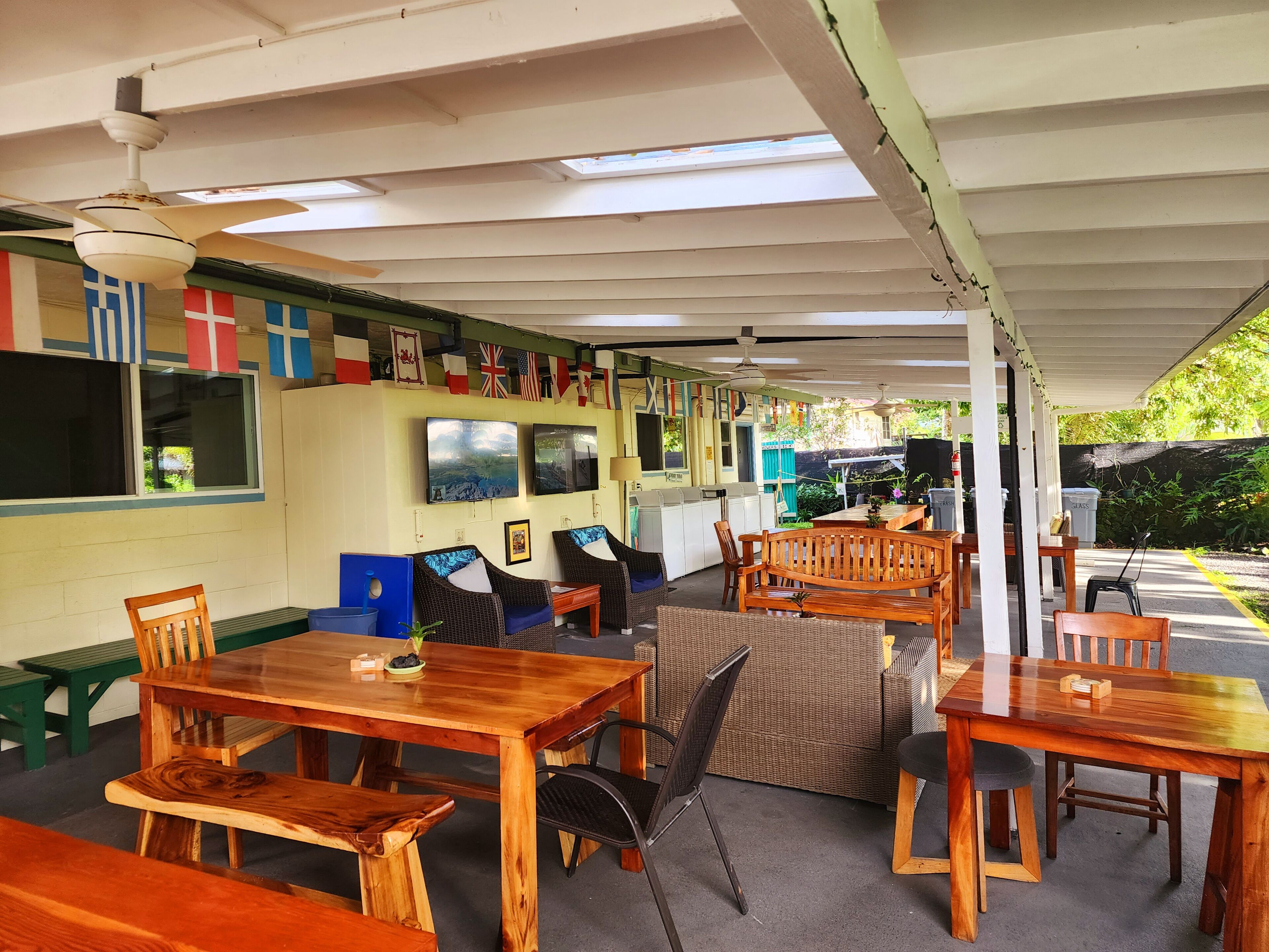Un espace extérieur abrité, avec des tables et des chaises en bois, un comptoir avec une caisse enregistreuse, et des drapeaux accrochés au mur.