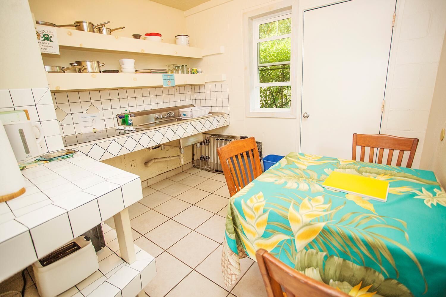 Una cucina compatta con piastrelle bianche, una finestra con vista su piante e un'area pranzo con tavolo coperto da una tovaglia a motivi tropicali.