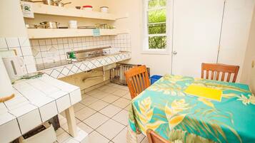 Una cucina compatta con piastrelle bianche, una finestra con vista su piante e un'area pranzo con tavolo coperto da una tovaglia a motivi tropicali.
