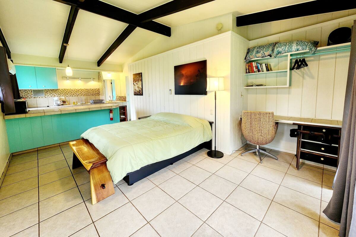 A bedroom with a bed, bedside table, and a desk with a chair. There is a kitchenette area with a sink and a mirror.