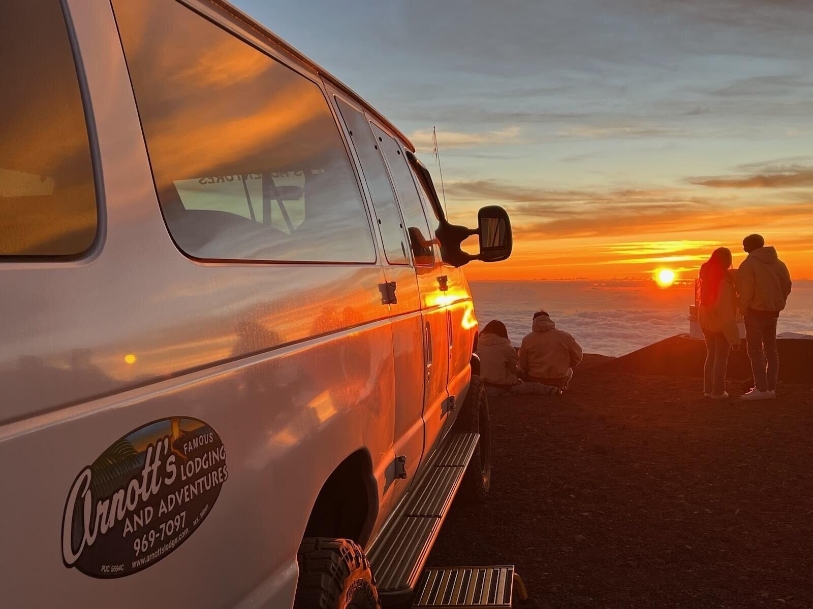 A white van with a logo and contact information, parked on a beach with a sunset in the background.