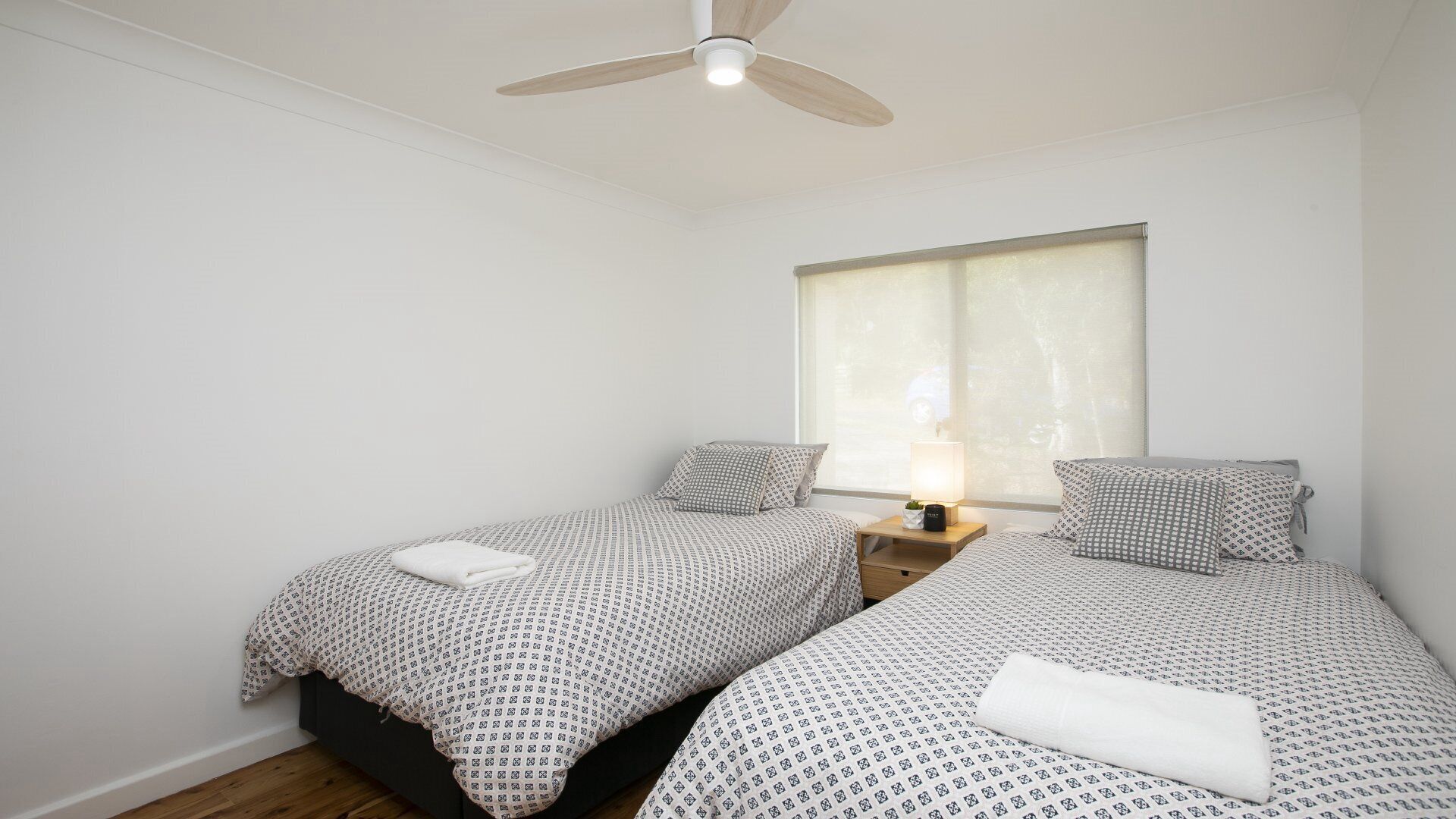 A bedroom with two beds, a ceiling fan, a window with blinds, and a bedside table with a lamp.
