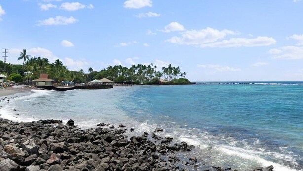A rocky beach with clear blue water, a small structure with a roof, and palm trees in the background. - Walk to Kahalu’u Beach | 2BR/2BA Condo with Lanai, Pool & A/C (Kailua-Kona)