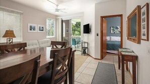 A spacious living area with a wooden dining table, a sofa, and a TV mounted on the wall. - Walk to Kahalu’u Beach | 2BR/2BA Condo with Lanai, Pool & A/C (Kailua-Kona)