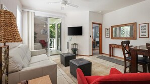A modern living room with a white sofa, a red armchair, a dining table with chairs, a flat-screen TV mounted on the wall, and a ceiling fan. - Walk to Kahalu’u Beach | 2BR/2BA Condo with Lanai, Pool & A/C (Kailua-Kona)