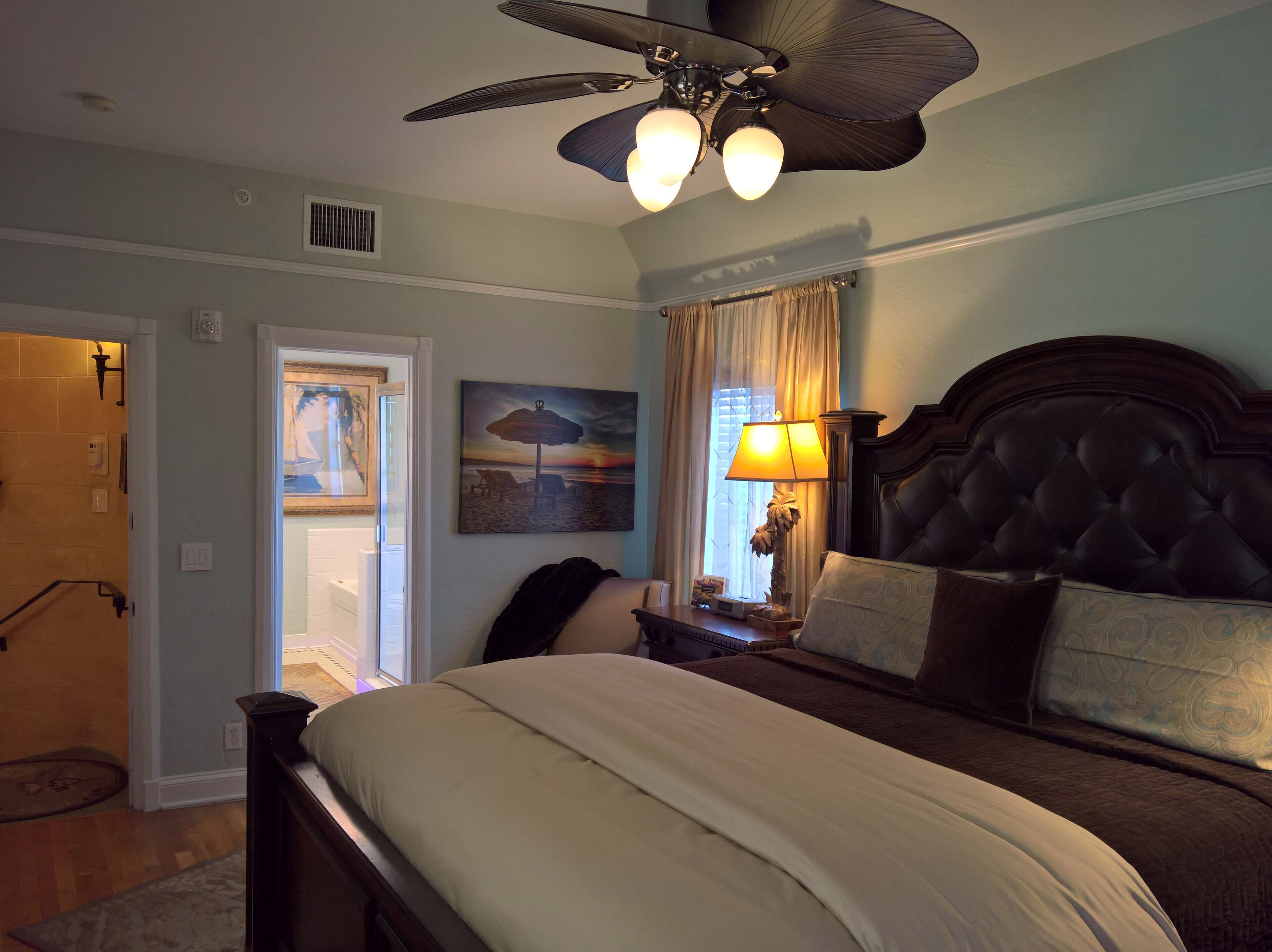 A bedroom with a large bed, a ceiling fan, a nightstand with a lamp, and a painting on the wall.