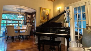 A grand piano in a spacious room with wooden floors, a dining area with a table and chairs, and a bookshelf filled with books. - Casa Grandview Inn (West Palm Beach)