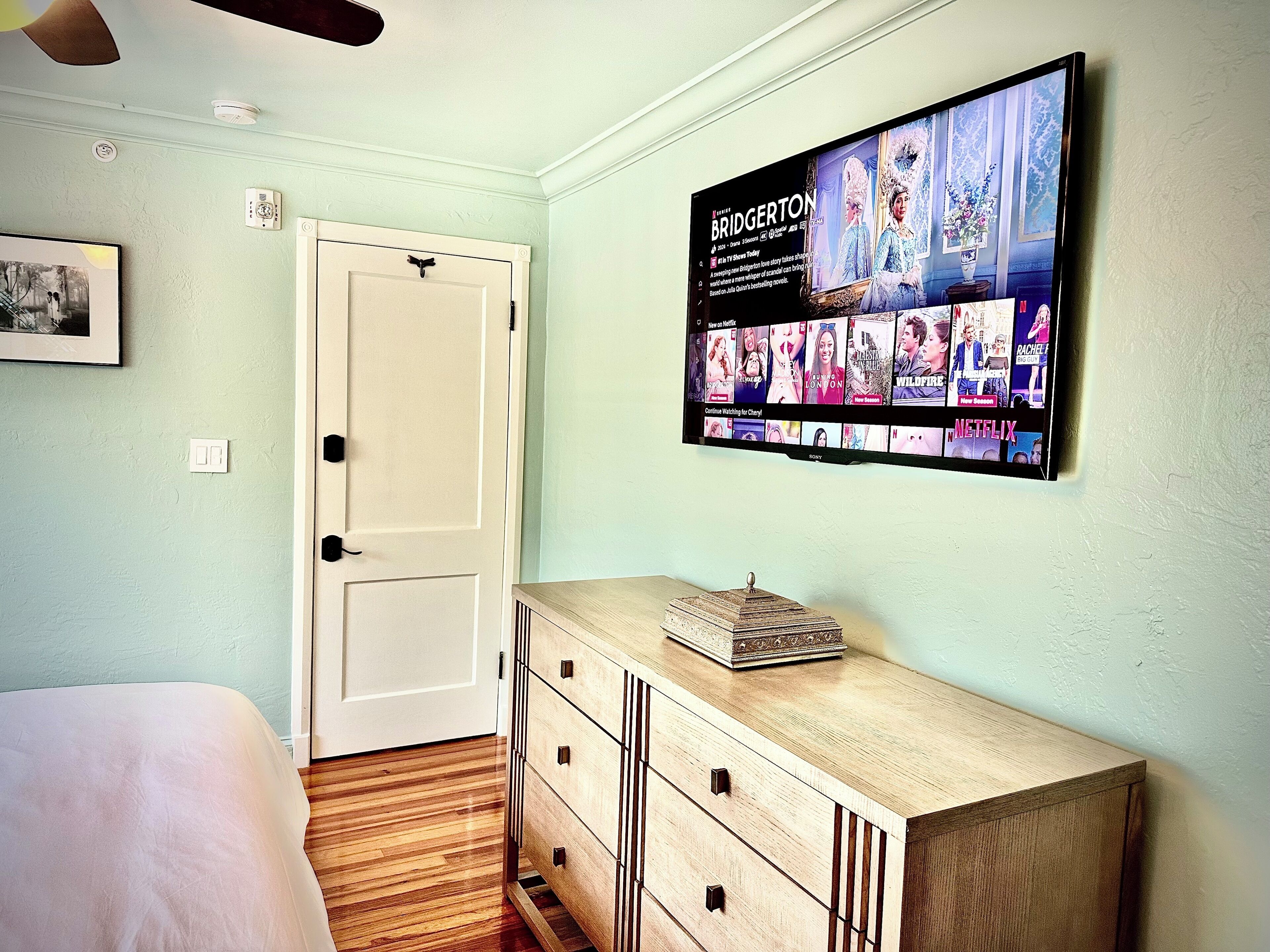 A bedroom with a bed, a dresser, a flat-screen TV mounted on the wall displaying a show, and a door.