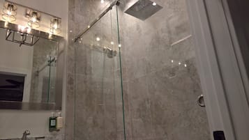 A modern bathroom with a glass-enclosed shower, marble walls, and a vanity with a mirror and two bottles of liquid soap.