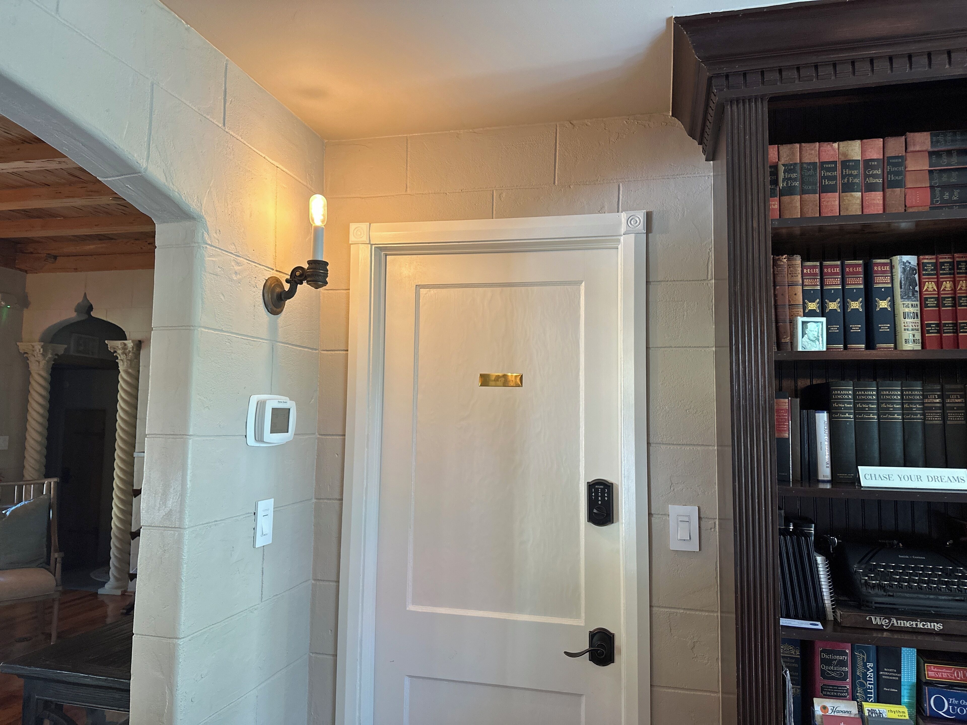 A room with a white door, a bookshelf with books, a thermostat, and a wall-mounted light fixture.