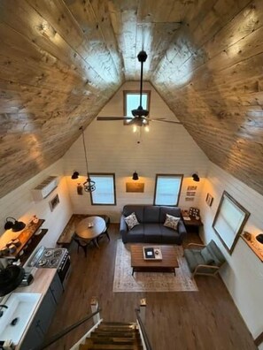 Een kamer in loftstijl met een schuine plafond, een plafondventilator, een bank, een salontafel, een eettafel en een keukenhoek.