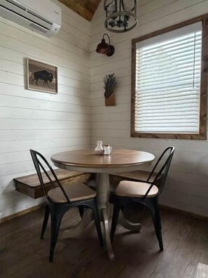 A dining area with a round table, two chairs, a wall-mounted air conditioner, a ceiling light fixture, a window with blinds, and a wall-mounted plant. - Black Hills Farmhouse, Guest House #1 (Custer)