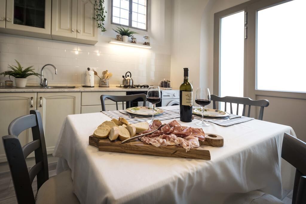 Un área de comedor con una mesa dispuesta para dos personas, que incluye una tabla de madera con diferentes carnes y quesos, una botella de vino y copas.