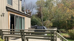 Eine Holzterrasse führt zu einem Gebäude mit einer Schiebetür, einem überdachten Grillplatz und Blick auf Bäume und Grünflächen.