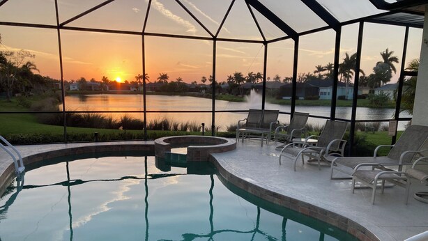 Une piscine intérieure offrant une vue sur un coucher de soleil se reflétant sur une étendue d’eau, entourée de chaises longues et d’un espace patio.