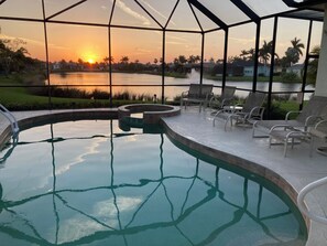 An indoor swimming pool with a view of a sunset over a body of water, surrounded by lounge chairs and a patio area. - Endless Summer, Gorgeous Sunsets! Spacious Lakefront 4/3 with pool and spa! (Fort Myers)