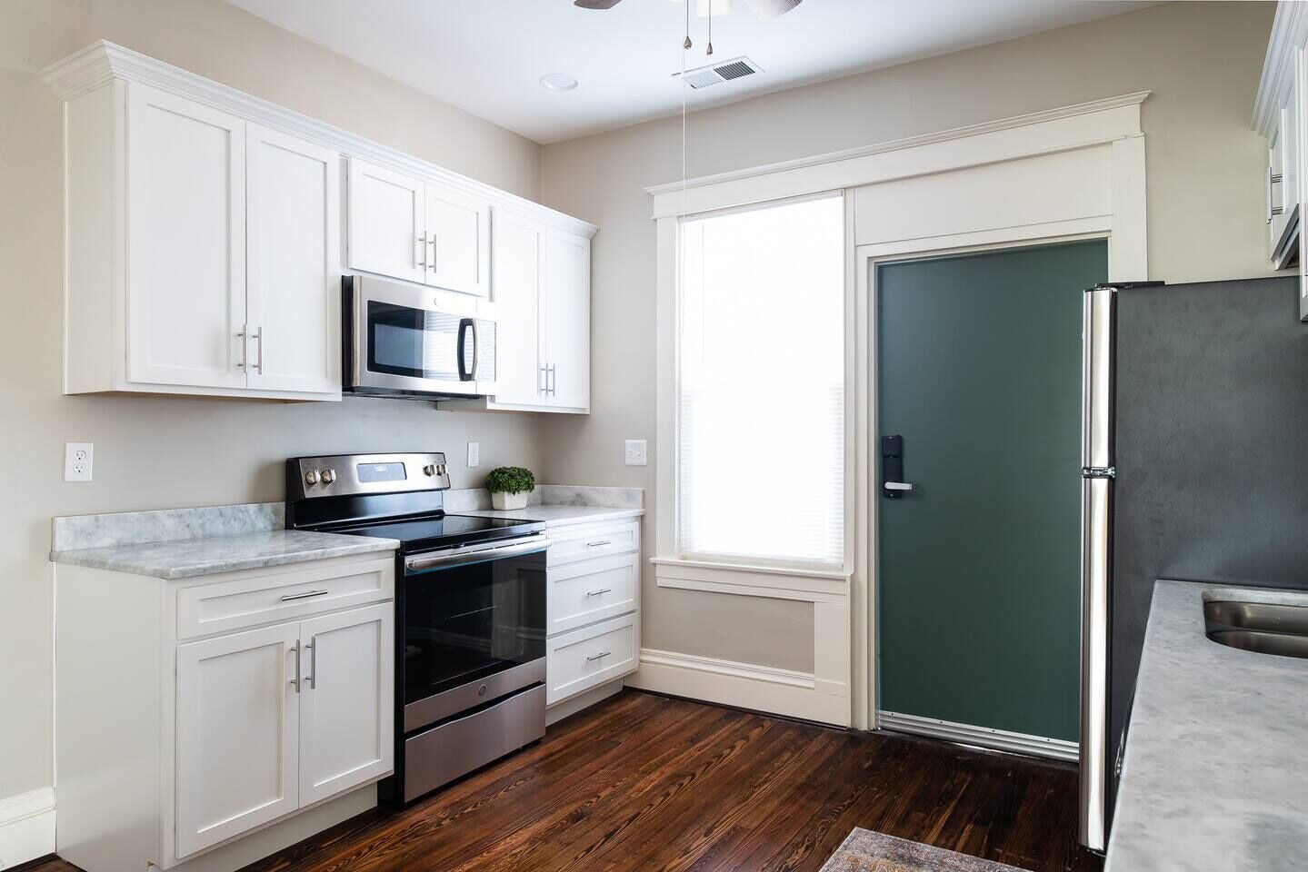 A kitchen with white cabinets, a stainless steel refrigerator, a microwave, a stove, and a window with blinds.