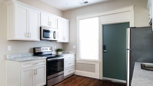 Una cocina con armarios blancos, un refrigerador de acero inoxidable, un microondas, una estufa y una ventana con persianas.