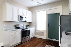A kitchen with white cabinets, a stainless steel refrigerator, a microwave, a stove, and a window with blinds. - Stylish Loft in Vibrant Neighborhood (Louisville)