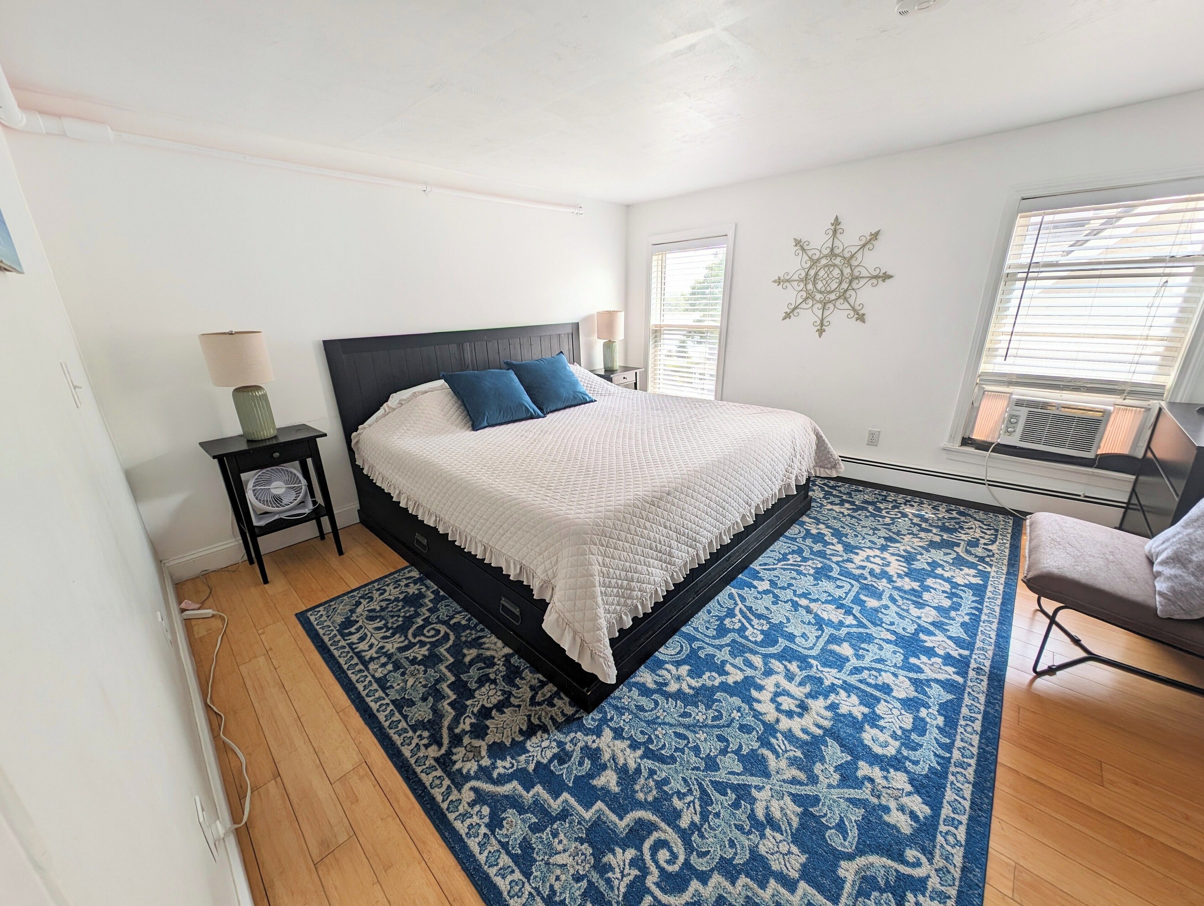 A bedroom with a bed, a nightstand, a lamp, a decorative wall piece, and a window with blinds.