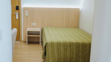 Ein kleines Zimmer mit einem Einzelbett, Holzboden, einem weißen Schreibtisch und einer grün gestreiften Bettdecke.