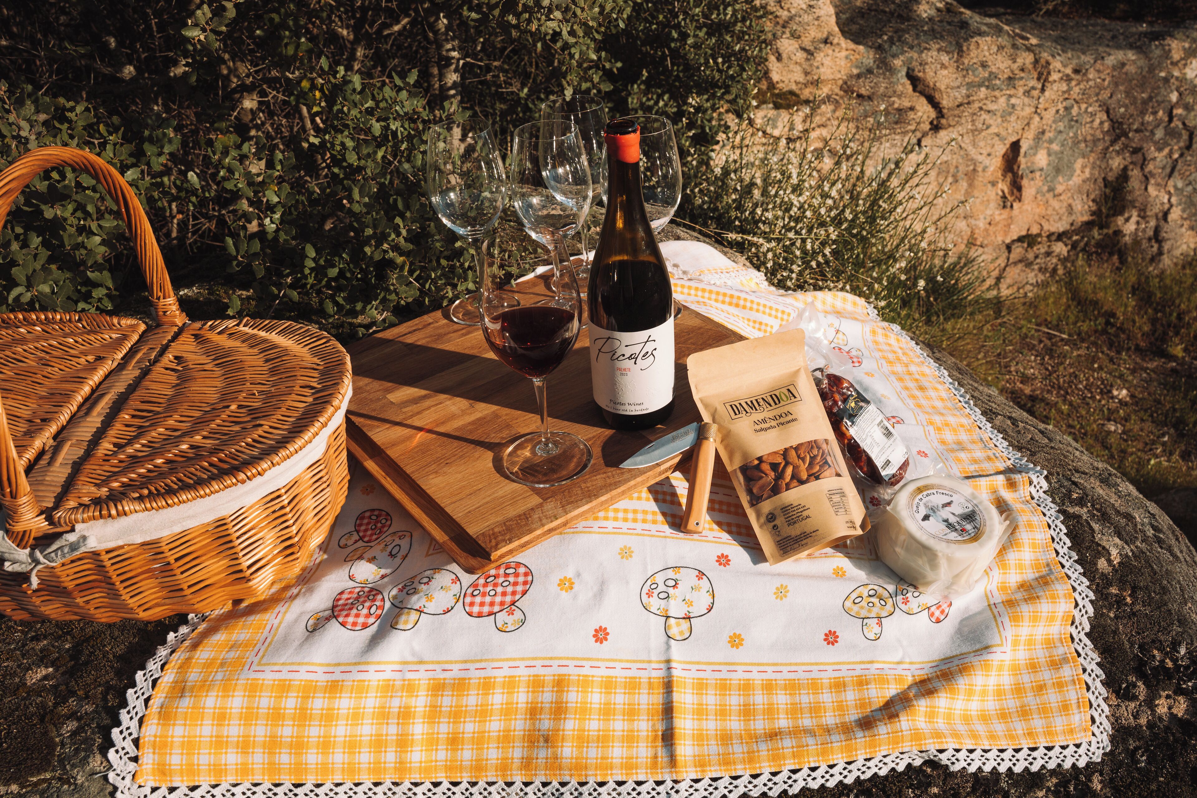 Un montaje para picnic con una cesta de mimbre, una botella de vino, copas de vino, un mantel a cuadros y una bolsa de aperitivos sobre una mesa de madera al aire libre.
