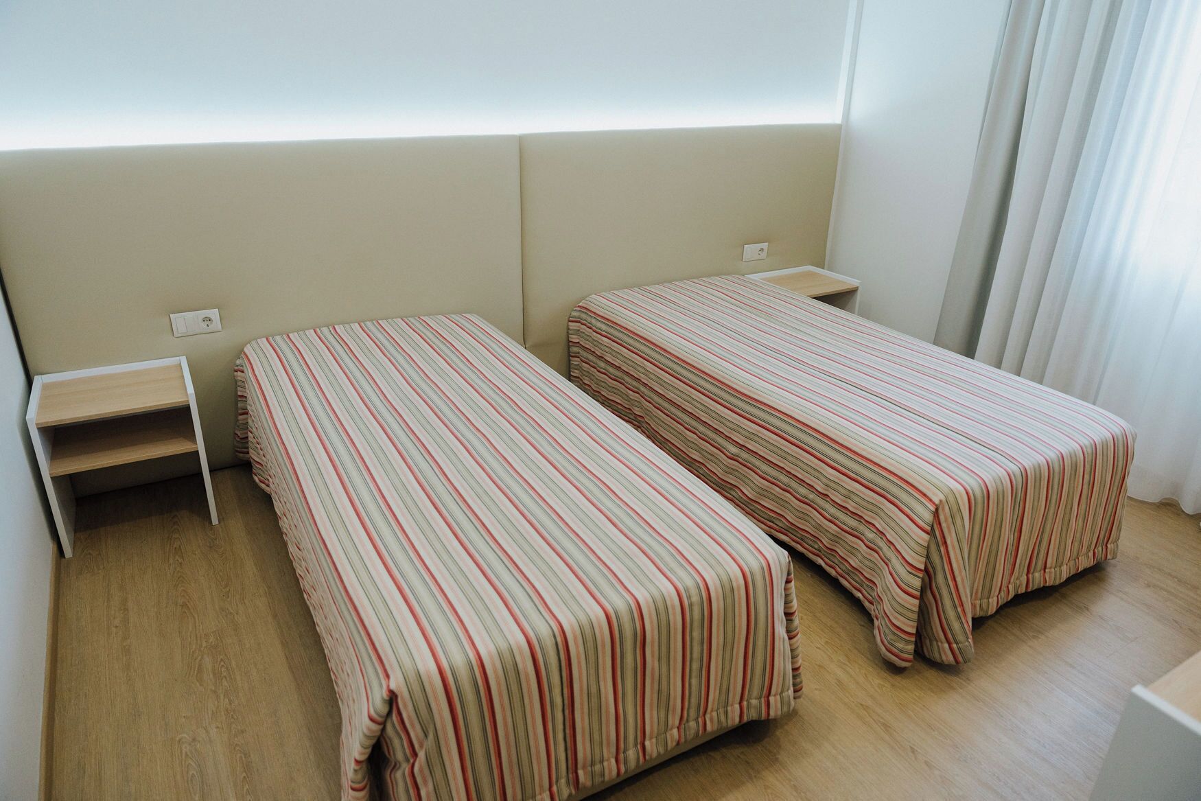 Une chambre avec deux lits simples, une petite table de chevet en bois et une prise électrique au mur.