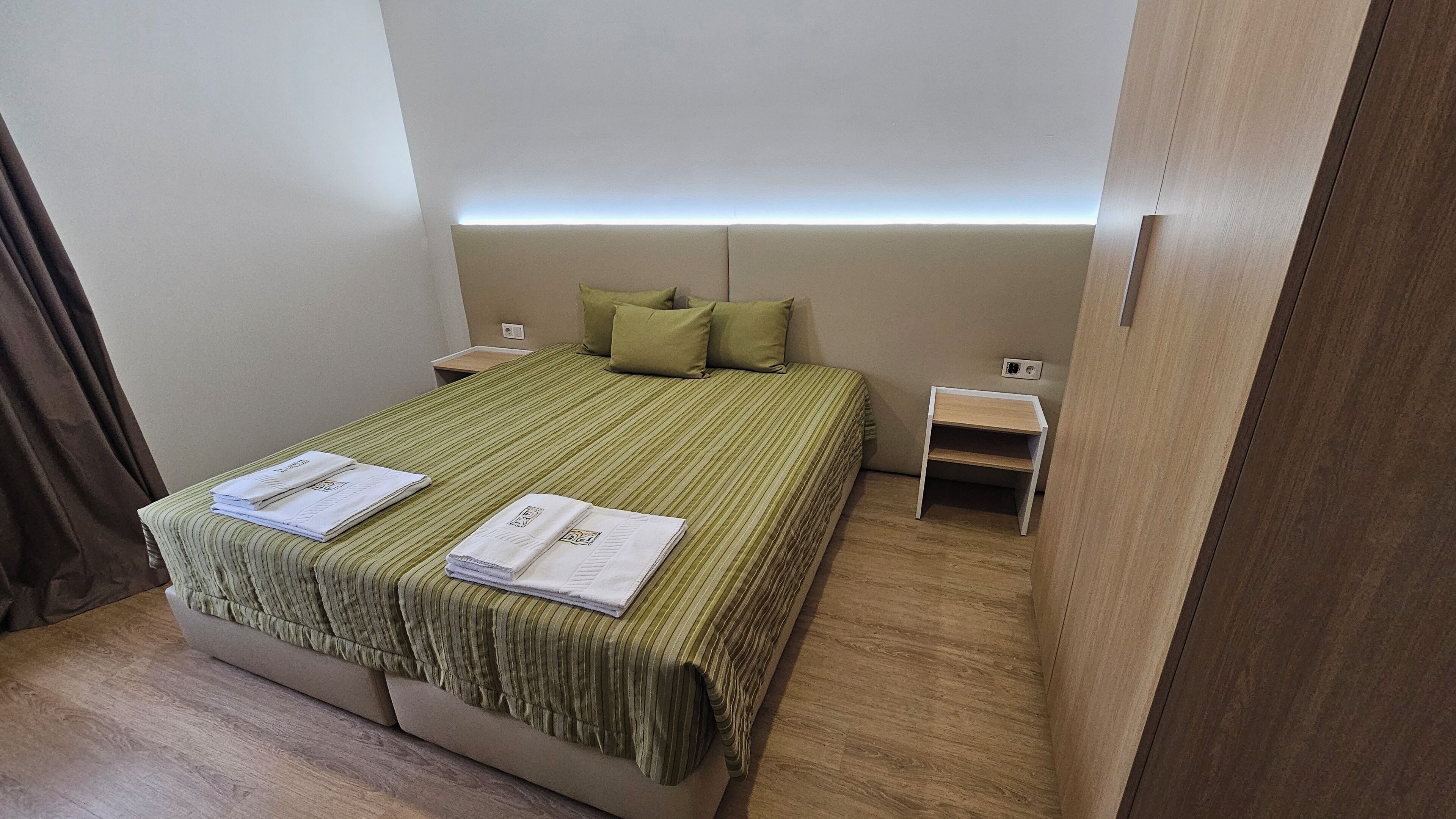 A neatly made bed with green bedding and two towels, a bedside table with a lamp, and a small wooden nightstand.