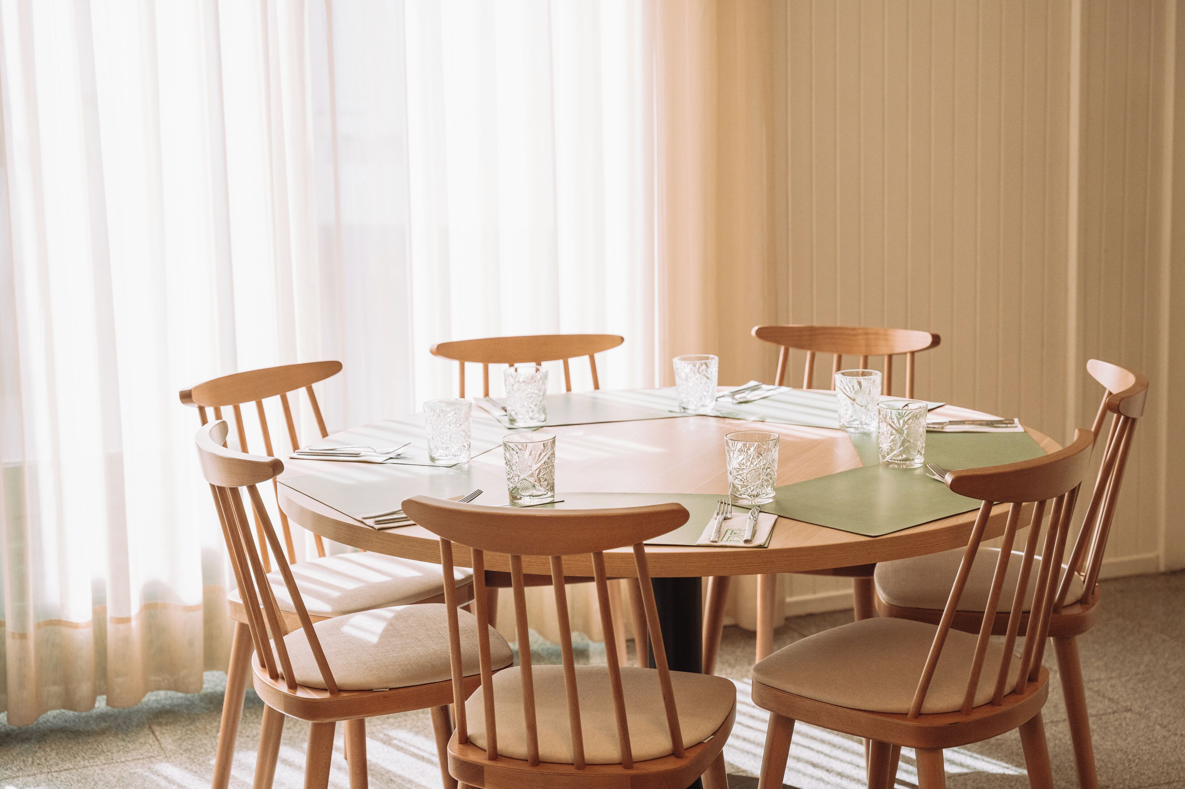 A round dining table with six wooden chairs, each with a cushioned seat. The table is set with clear glassware and a green placemat.