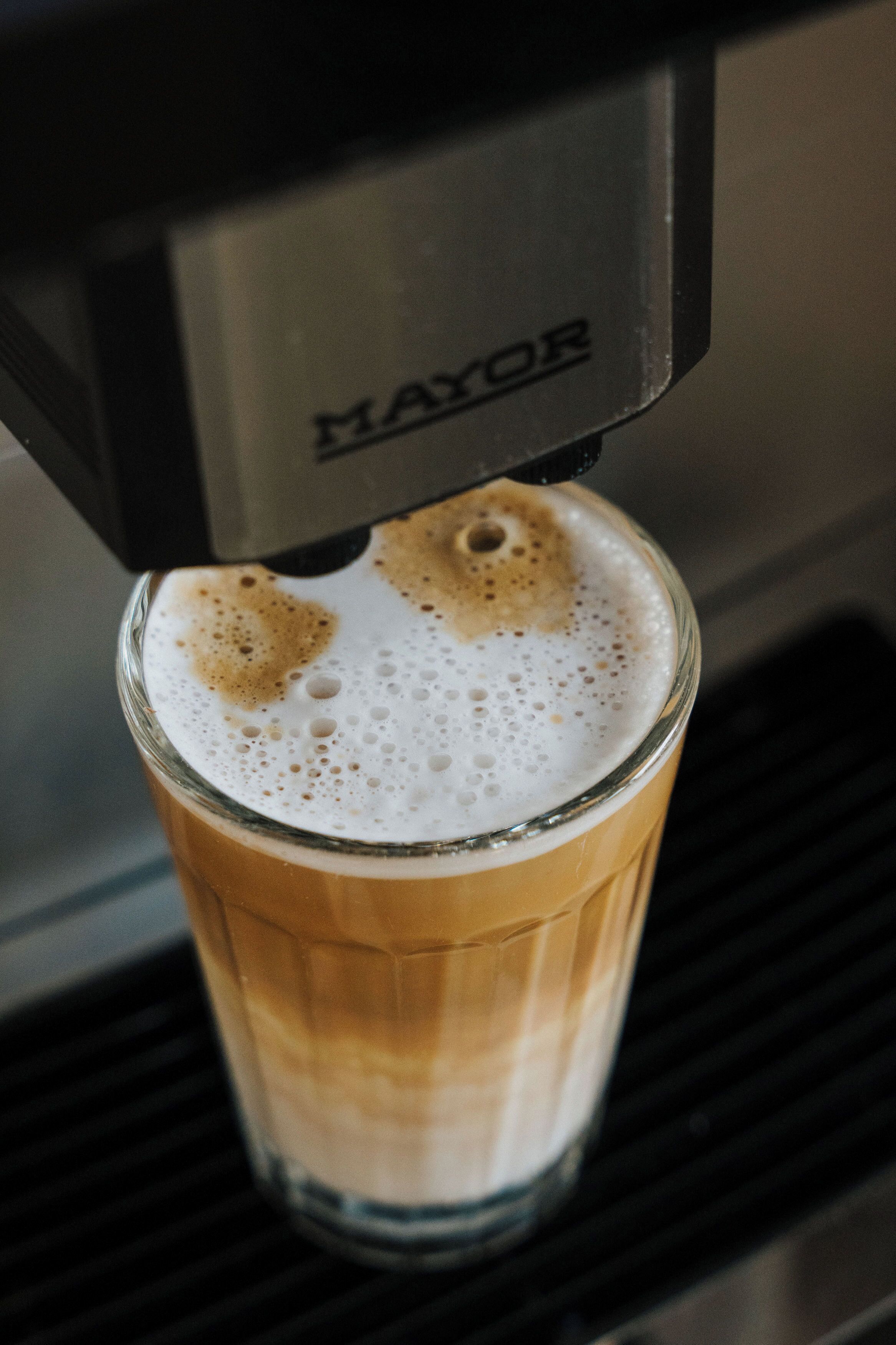 A glass of coffee with foam on top, being dispensed from a machine labeled "MAYOR".