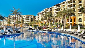 Zona de piscina en un resort, con múltiples sillas de descanso y sombrillas, rodeada de palmeras y un edificio de varios pisos con balcones.