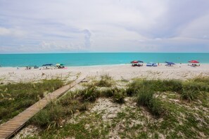 Una playa con un paseo marítimo de madera que llega hasta la orilla, rodeada de arena y vegetación escasa.