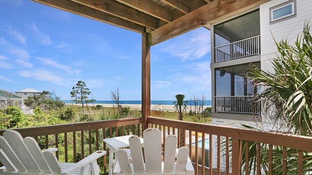 Eine Holzterrasse mit weißen Adirondack-Stühlen mit Blick auf einen Strand und ein zweistöckiges Gebäude mit Balkonen.