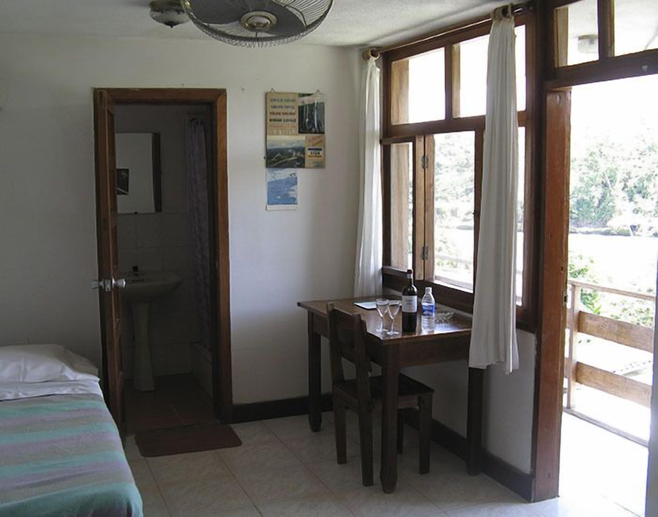 A room with a bed, a desk with chairs, a window with curtains, a fan, and a poster on the wall.