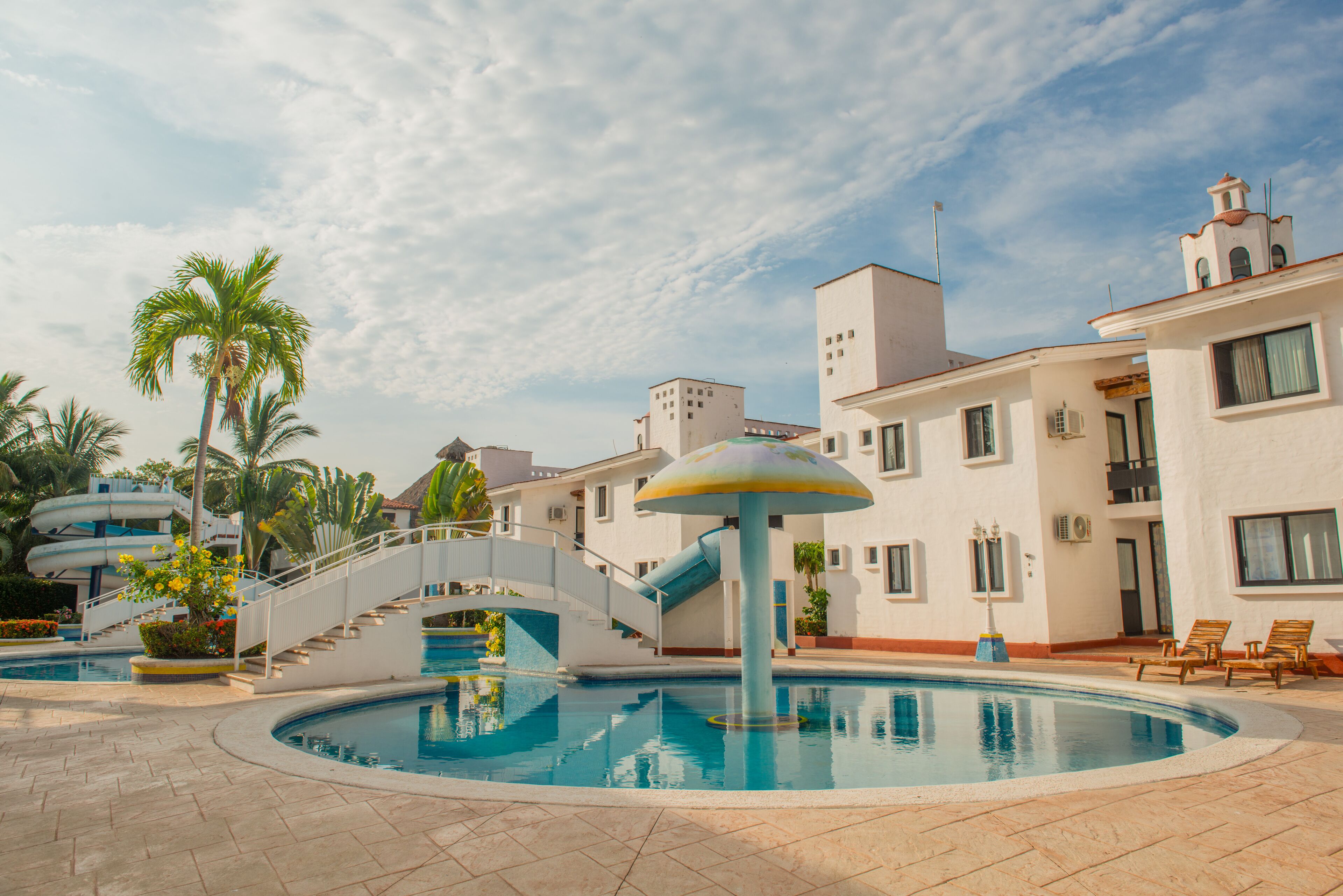 Uma piscina com uma fonte central, cercada por palmeiras e um edifício branco com varandas.