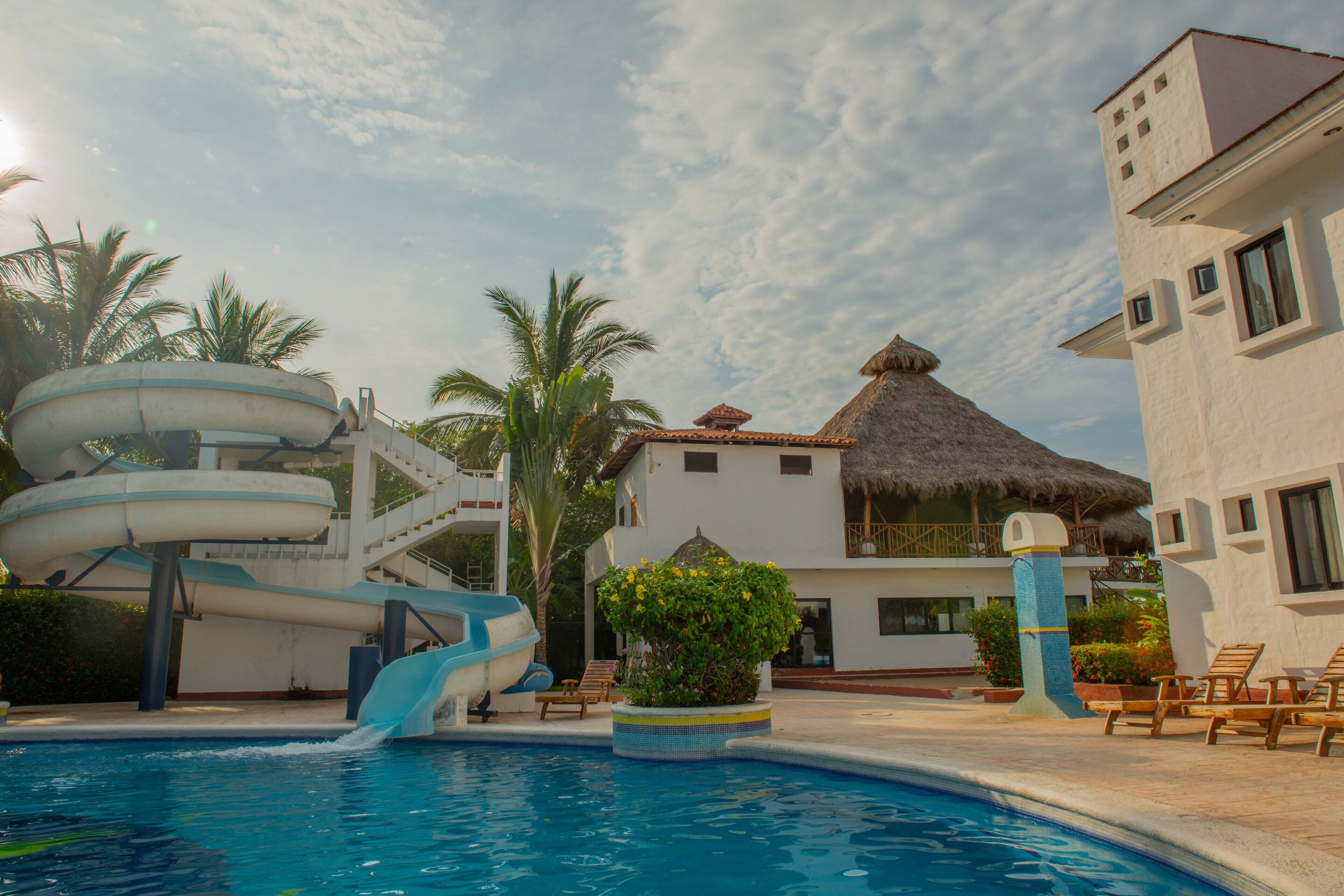 A swimming pool with a water slide, palm trees, and a building with a thatched roof.