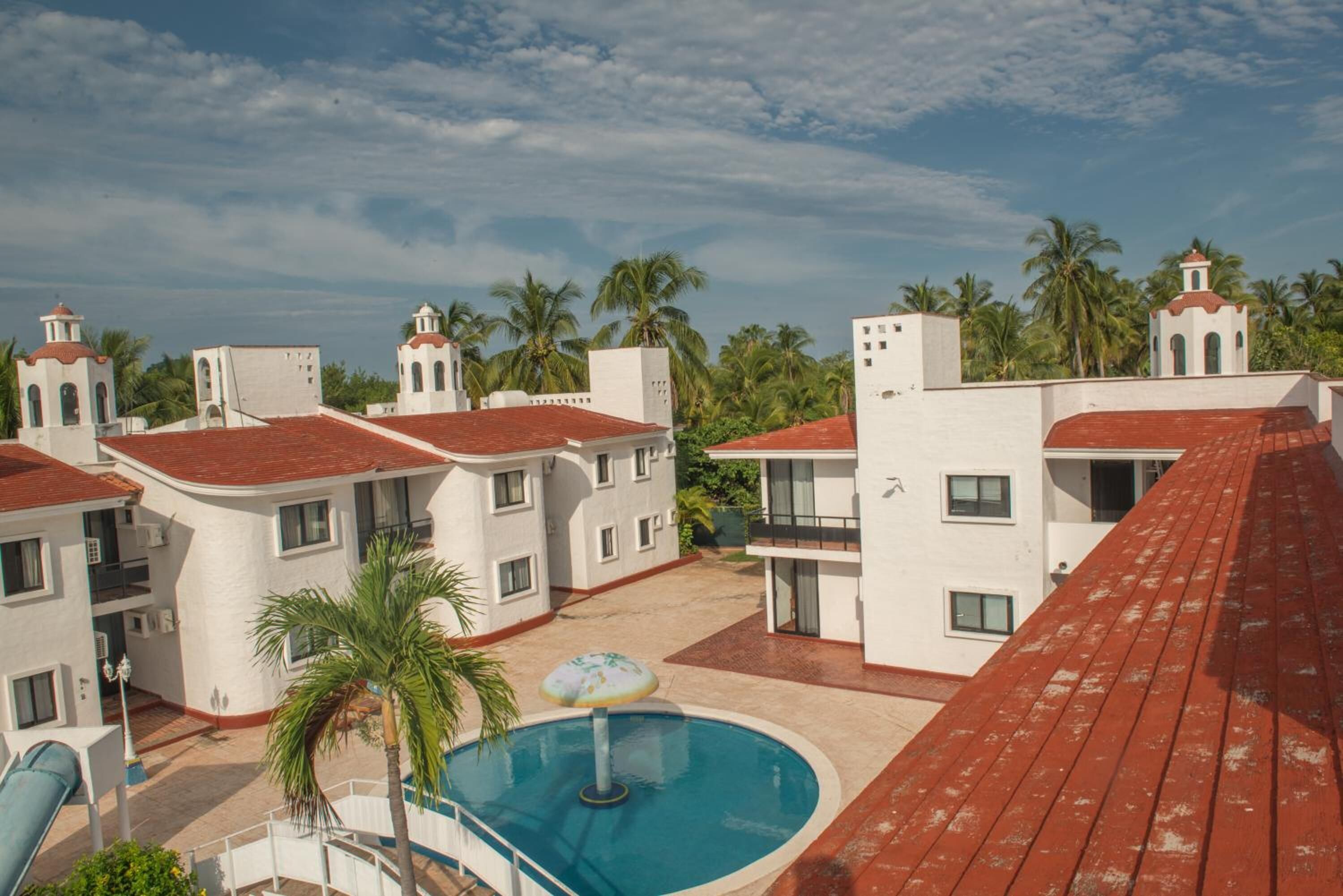 A resort with multiple white buildings, red roofs, and a swimming pool with a palm tree and umbrella.
