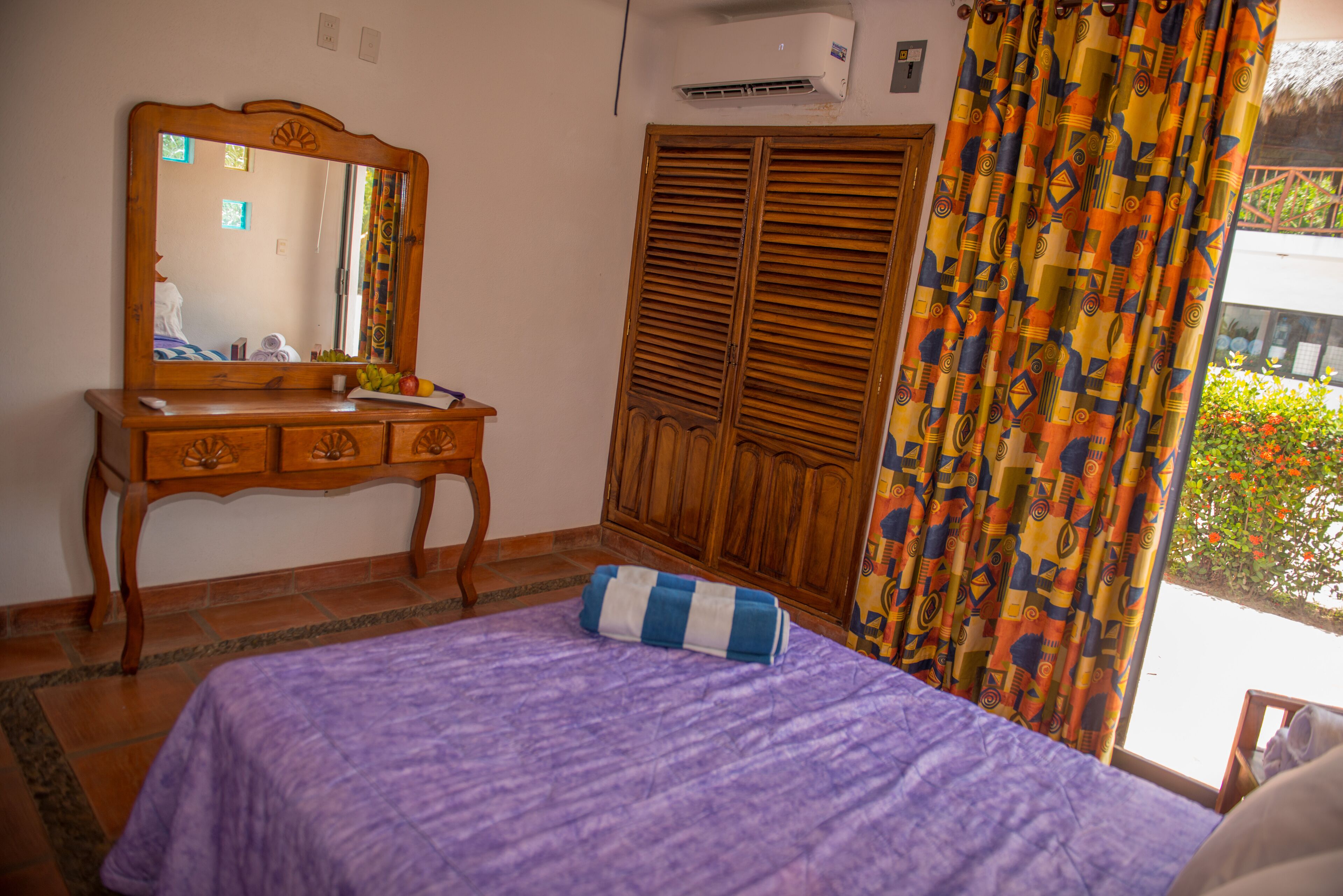 Un dormitorio con un aparador de madera, una cama con colcha púrpura, un espejo, un armario y una ventana con cortinas.