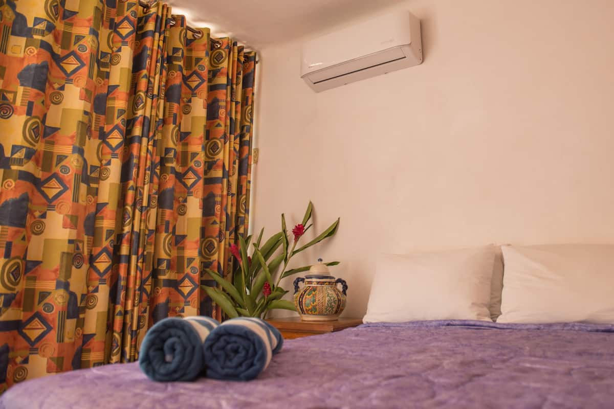 A bedroom with a bed, pillows, a rolled-up towel, a vase with flowers, and a wall-mounted air conditioner.