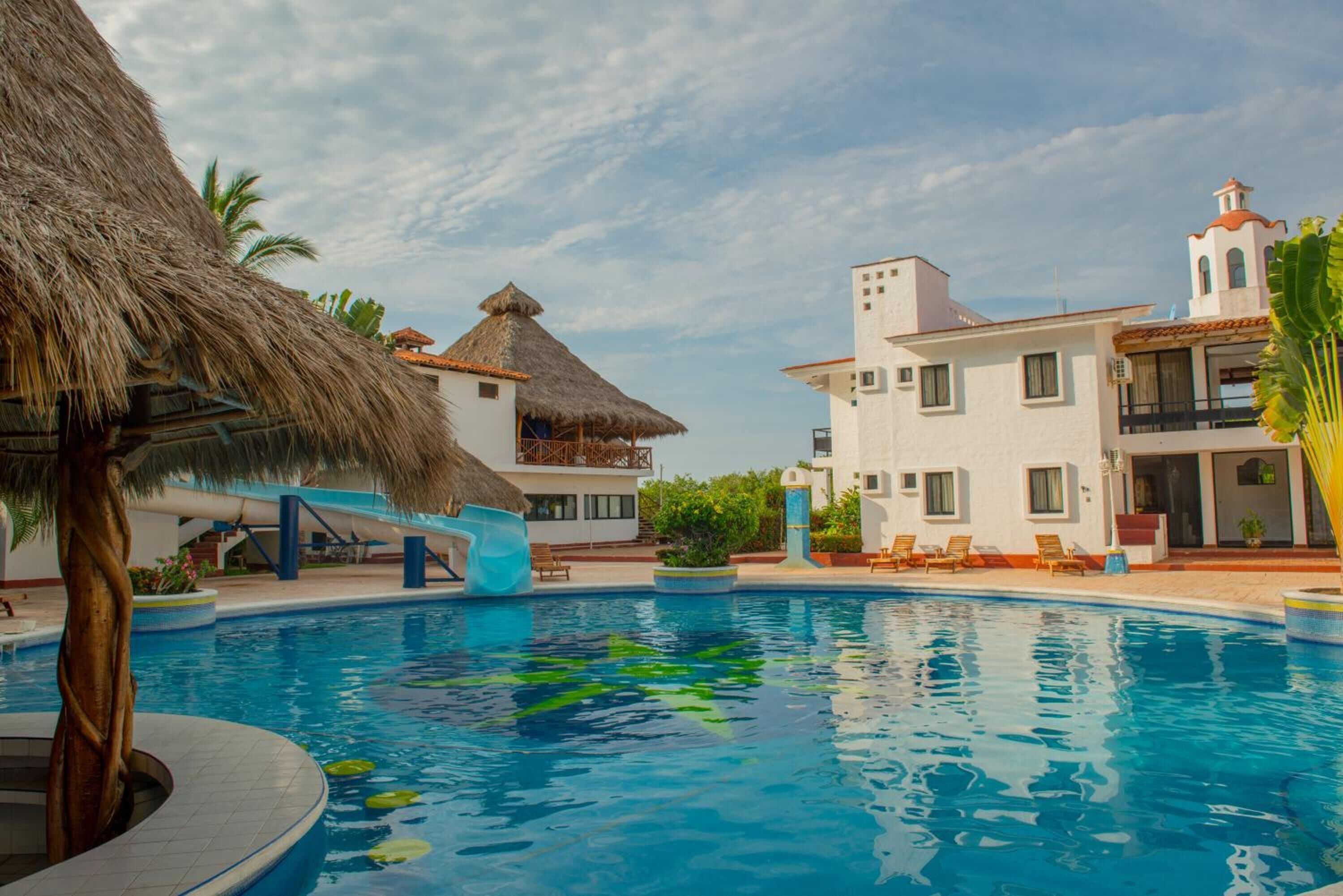 A swimming pool with a thatched roof structure, white buildings, and a clear sky.