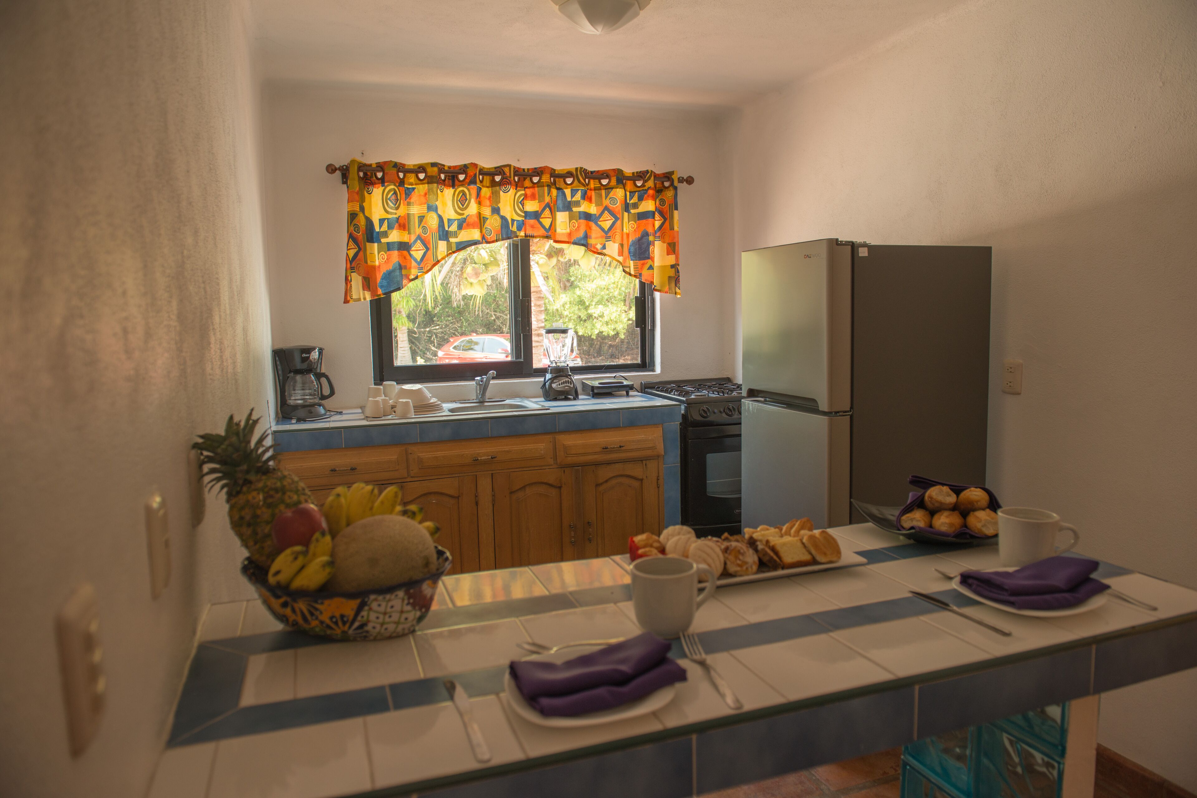 A kitchen with a refrigerator, stove, and window with curtains. There is a fruit bowl on the counter and a plate of pastries.